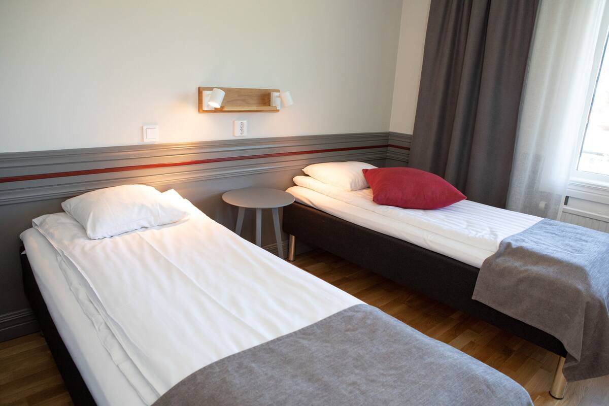Une chambre d’hôtel avec deux lits simples, une petite table ronde, une étagère avec des articles de toilette et une fenêtre avec des rideaux.