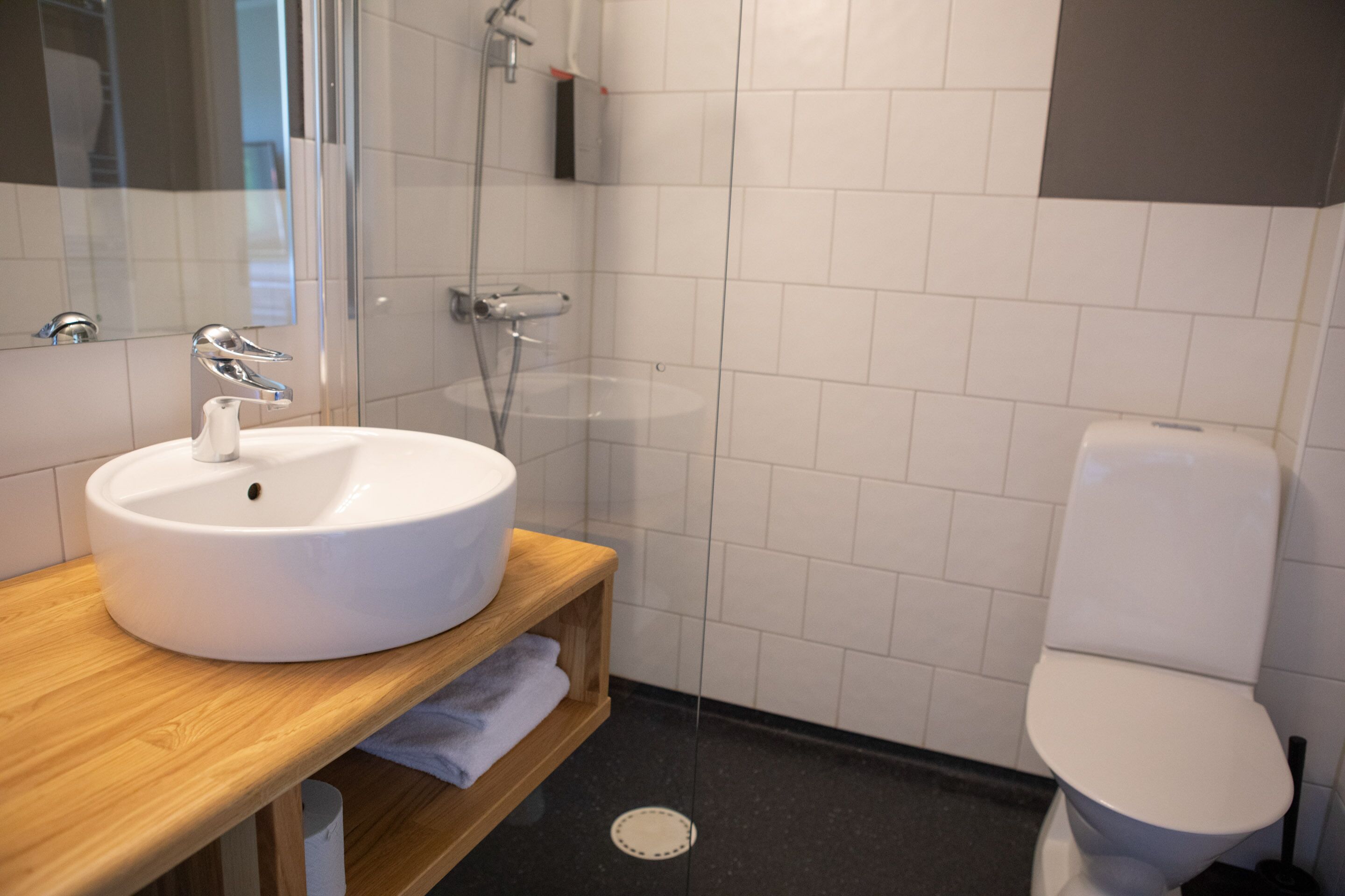 Un baño moderno con un lavabo blanco, un mueble de tocador de madera, una cabina de ducha de vidrio y un inodoro blanco.