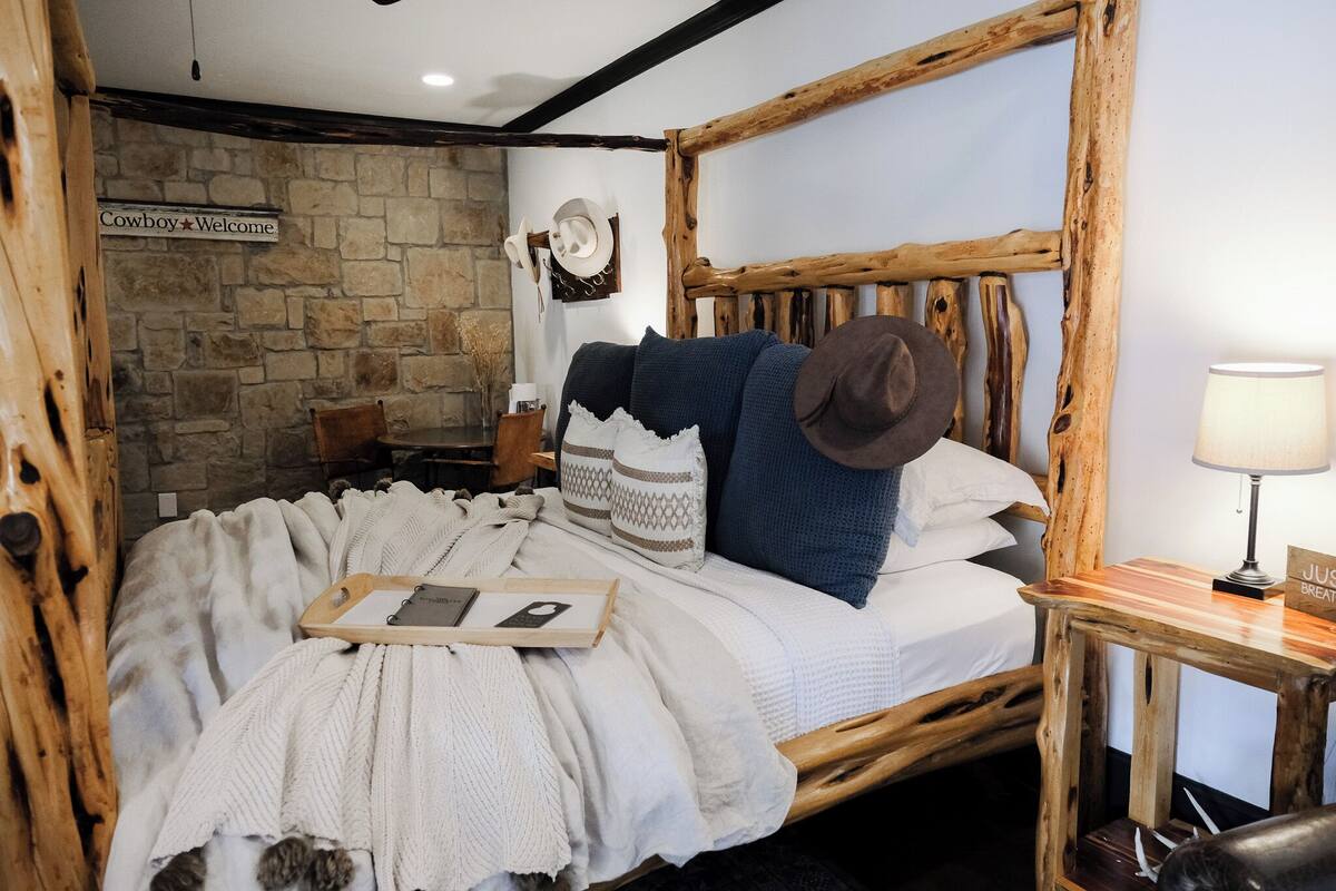Une chambre rustique avec un mur en pierre, un cadre de lit en bois, une pancarte de bienvenue, un chapeau et une table de chevet avec une lampe.