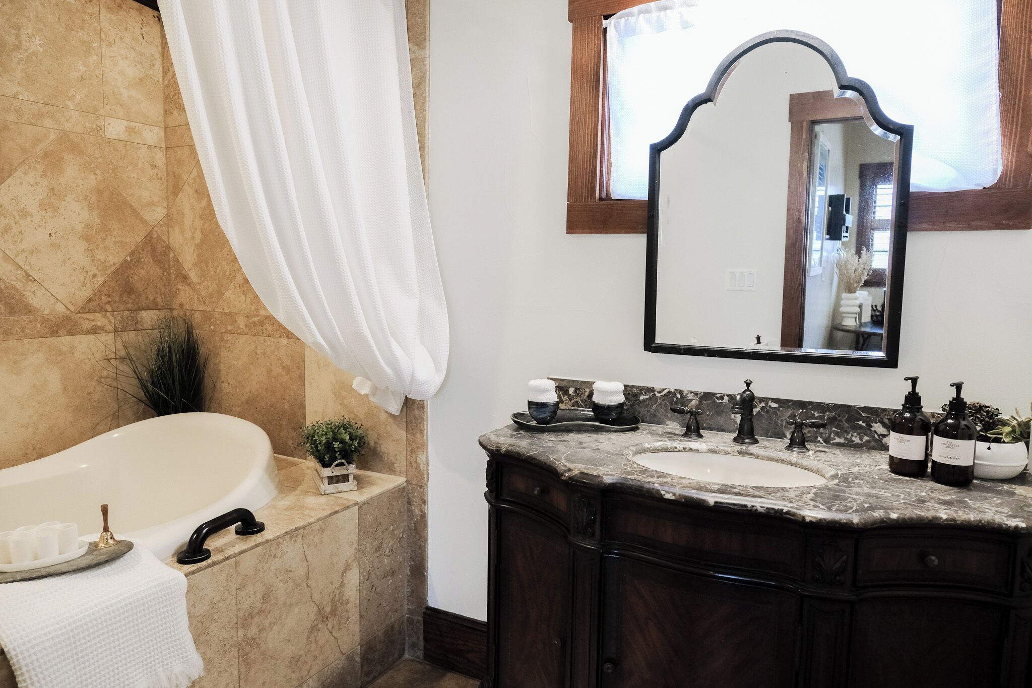 Une salle de bain avec une baignoire, deux lavabos, un meuble-lavabo avec des armoires en bois foncé, un grand miroir et une fenêtre avec un rideau.