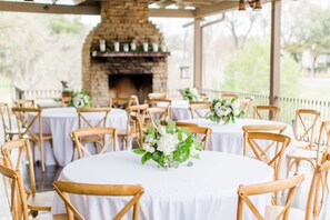 A covered outdoor dining area with round tables and wooden chairs, a stone fireplace, and a view of greenery.