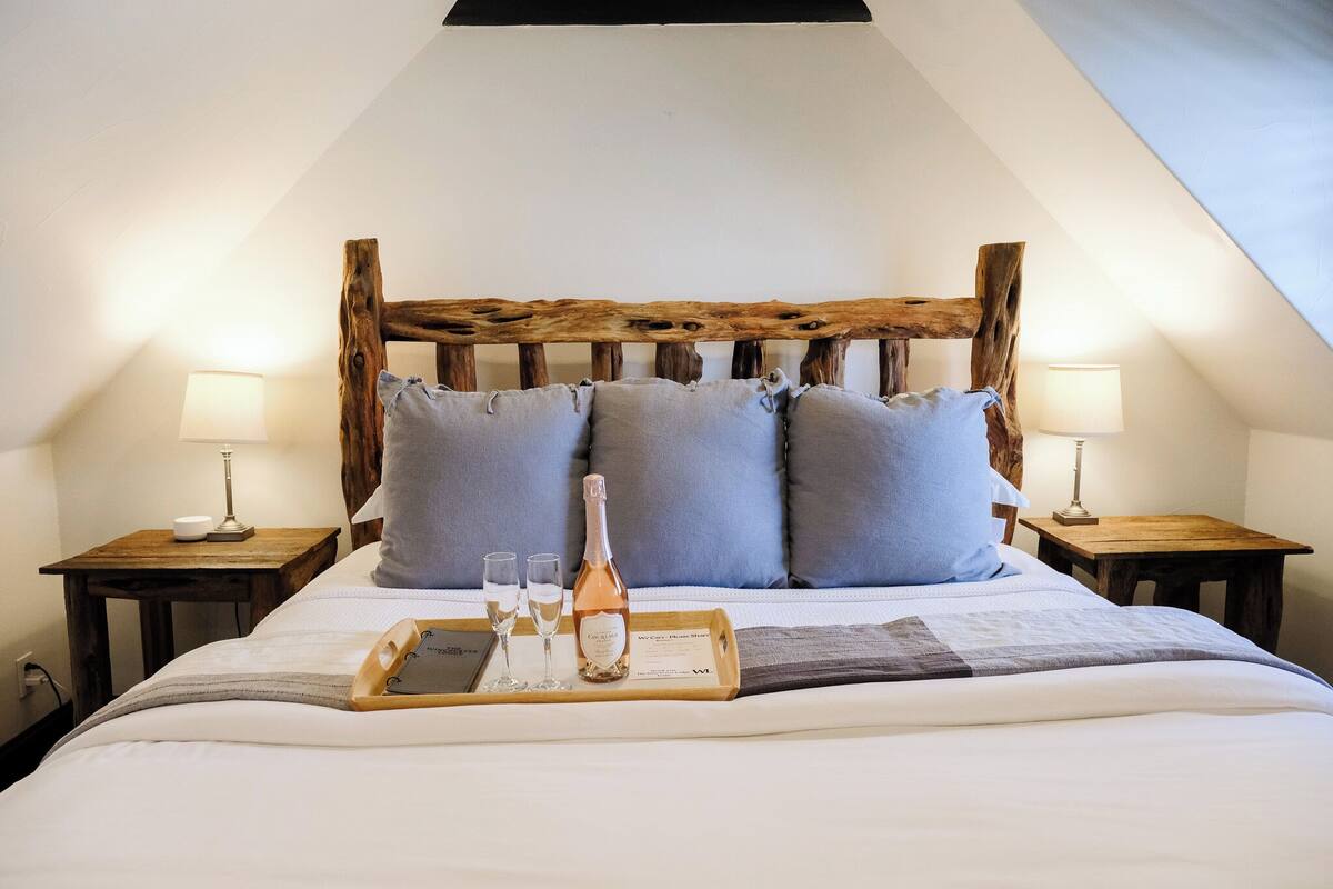 Un lit bien fait, avec une tête de lit en bois, deux tables de chevet avec des lampes, un plateau avec une bouteille et des verres, et un oreiller sur le lit.