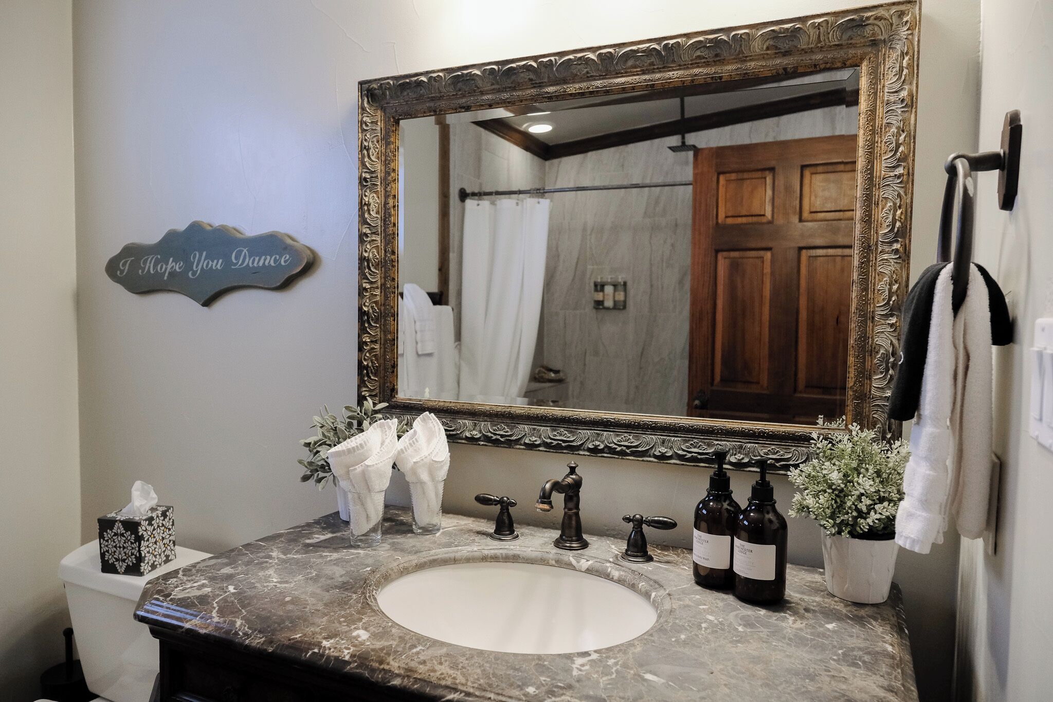 A bathroom with a marble sink, a large ornate mirror, a towel rack, a potted plant, and a sign that says "I Hope You Dance".