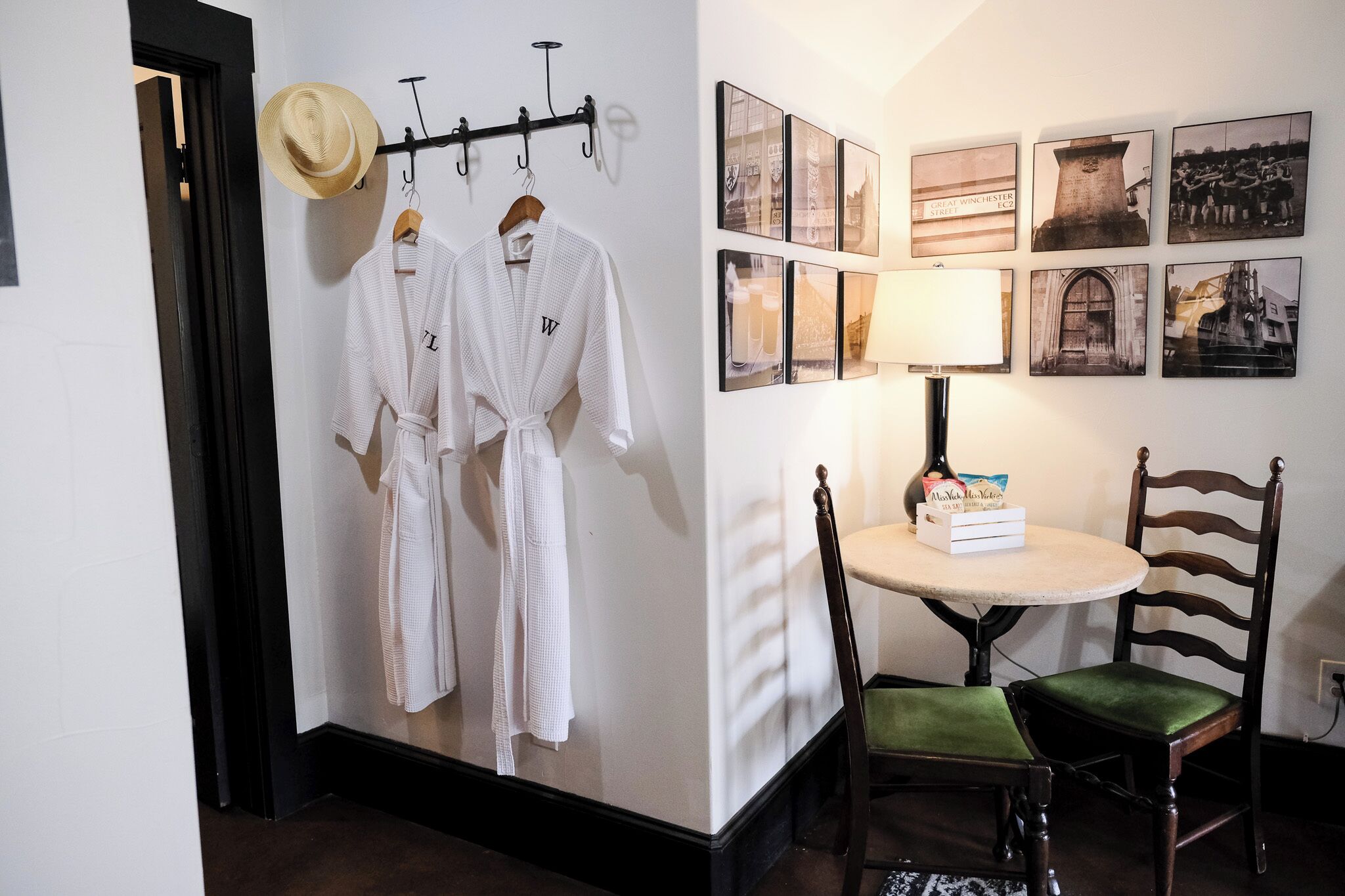 A room with a wall-mounted robe rack holding two white robes, a round table with a lamp and a box, and a wooden chair.