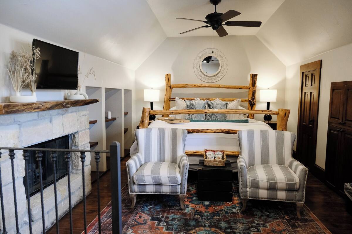 Une chambre avec une cheminée en pierre, un lit avec une tête de lit en bois, deux fauteuils rembourrés, un ventilateur de plafond et un téléviseur fixé au mur.