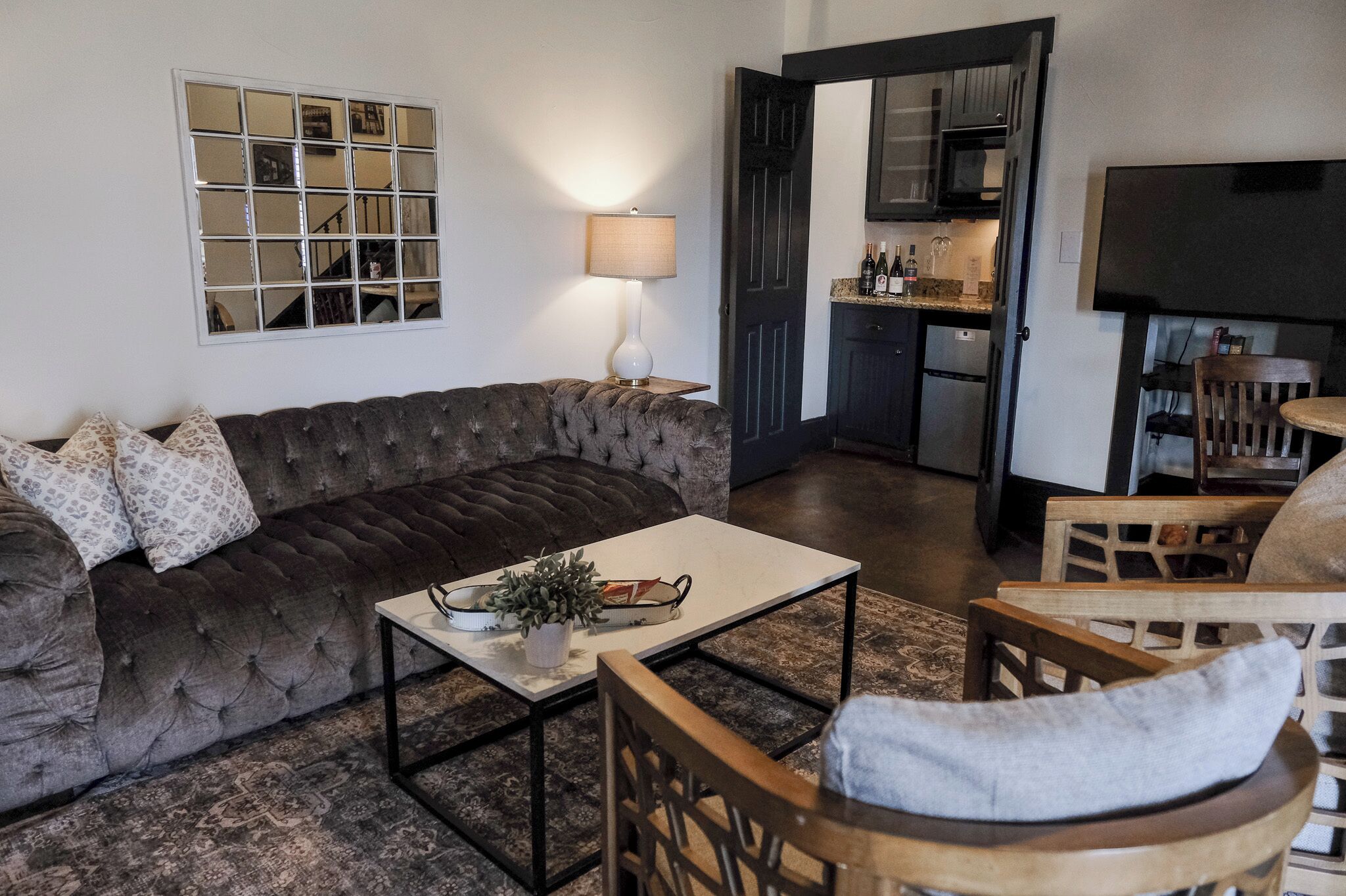 A living room with a dark brown sofa, a white coffee table, and a wooden chair. A lamp is on the table, and a kitchen is visible through an open door.