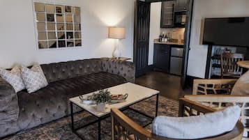 A living room with a dark brown sofa, a white coffee table, and a wooden chair. A lamp is on the table, and a kitchen is visible through an open door.