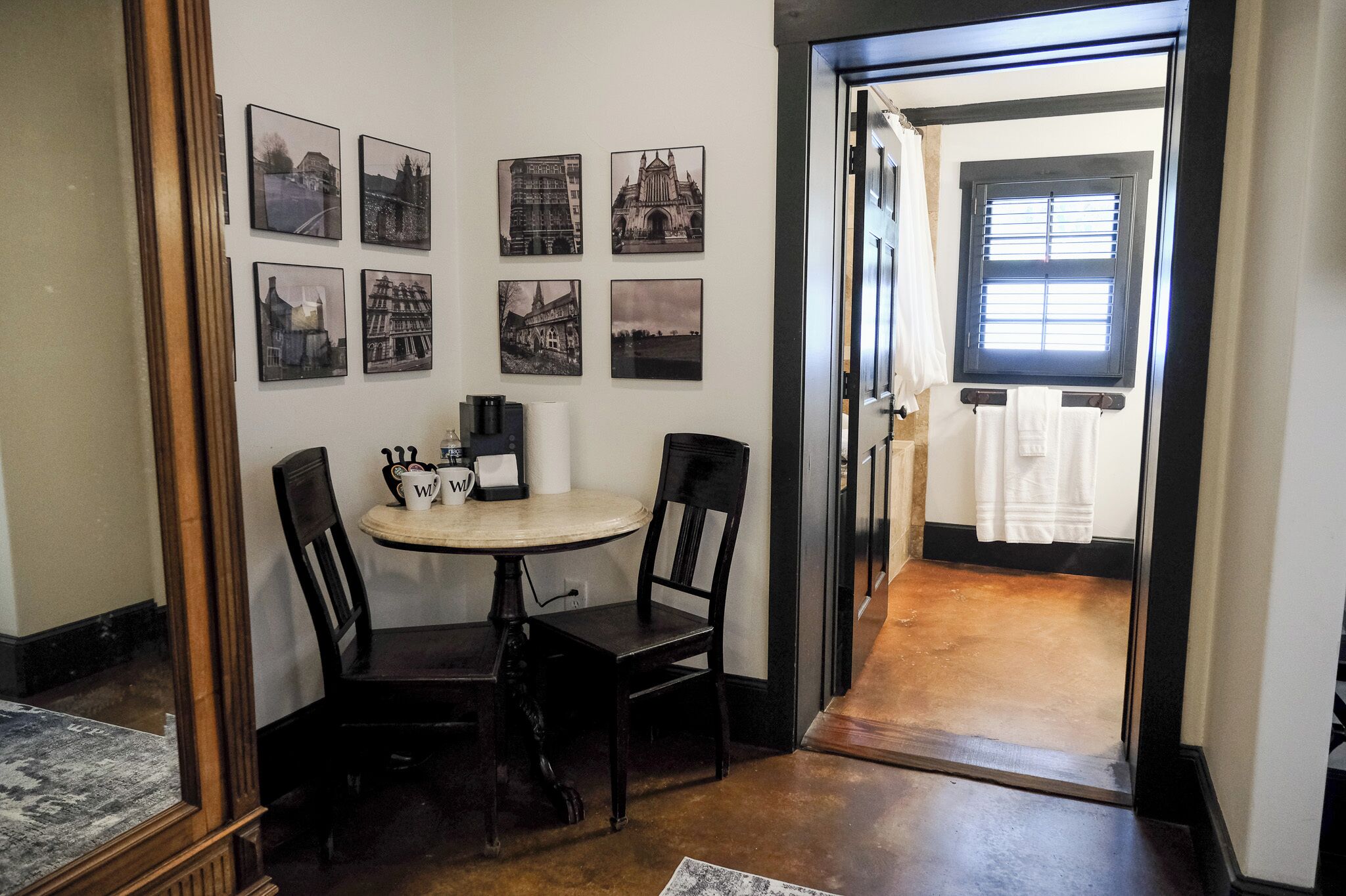 A dining area with a round table and four chairs, a wall with six black and white photographs, and a doorway leading to a room with a window and a towel rack.