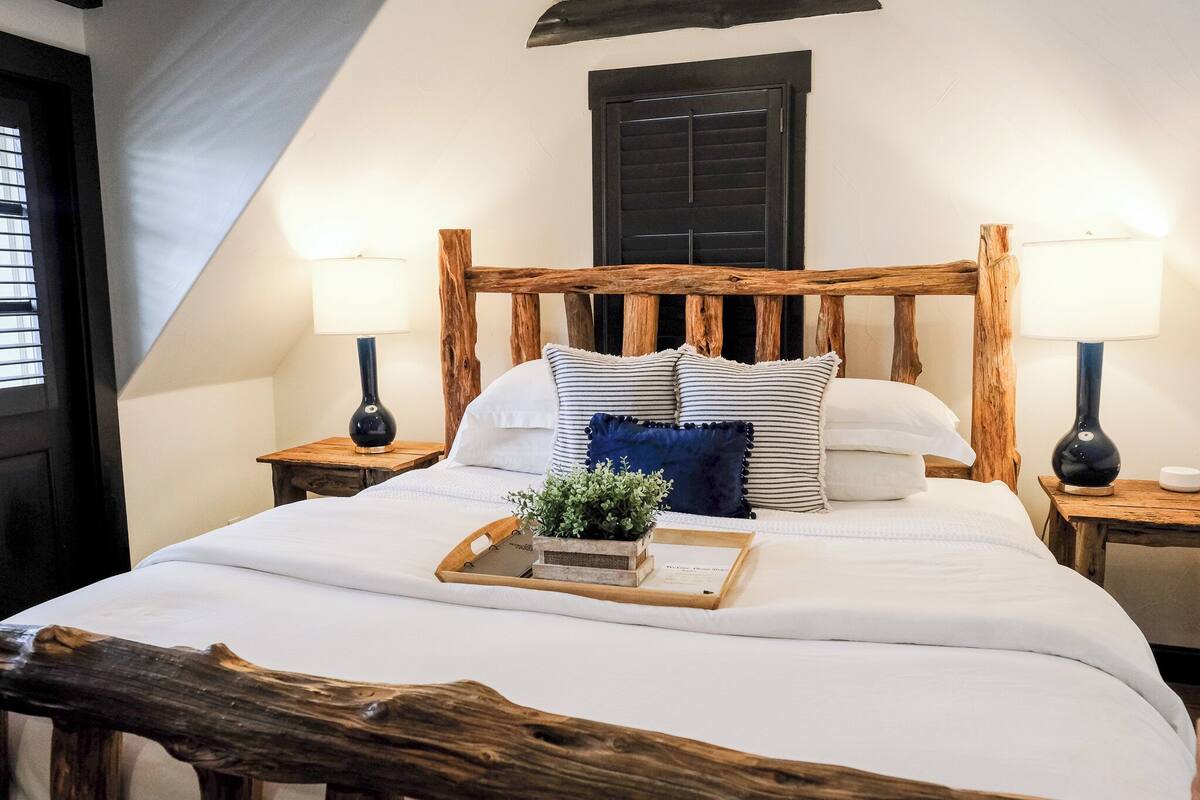Une chambre à coucher avec une tête de lit en bois, du linge de lit blanc, deux lampes de chevet, un plateau avec une plante et une fenêtre avec des volets.
