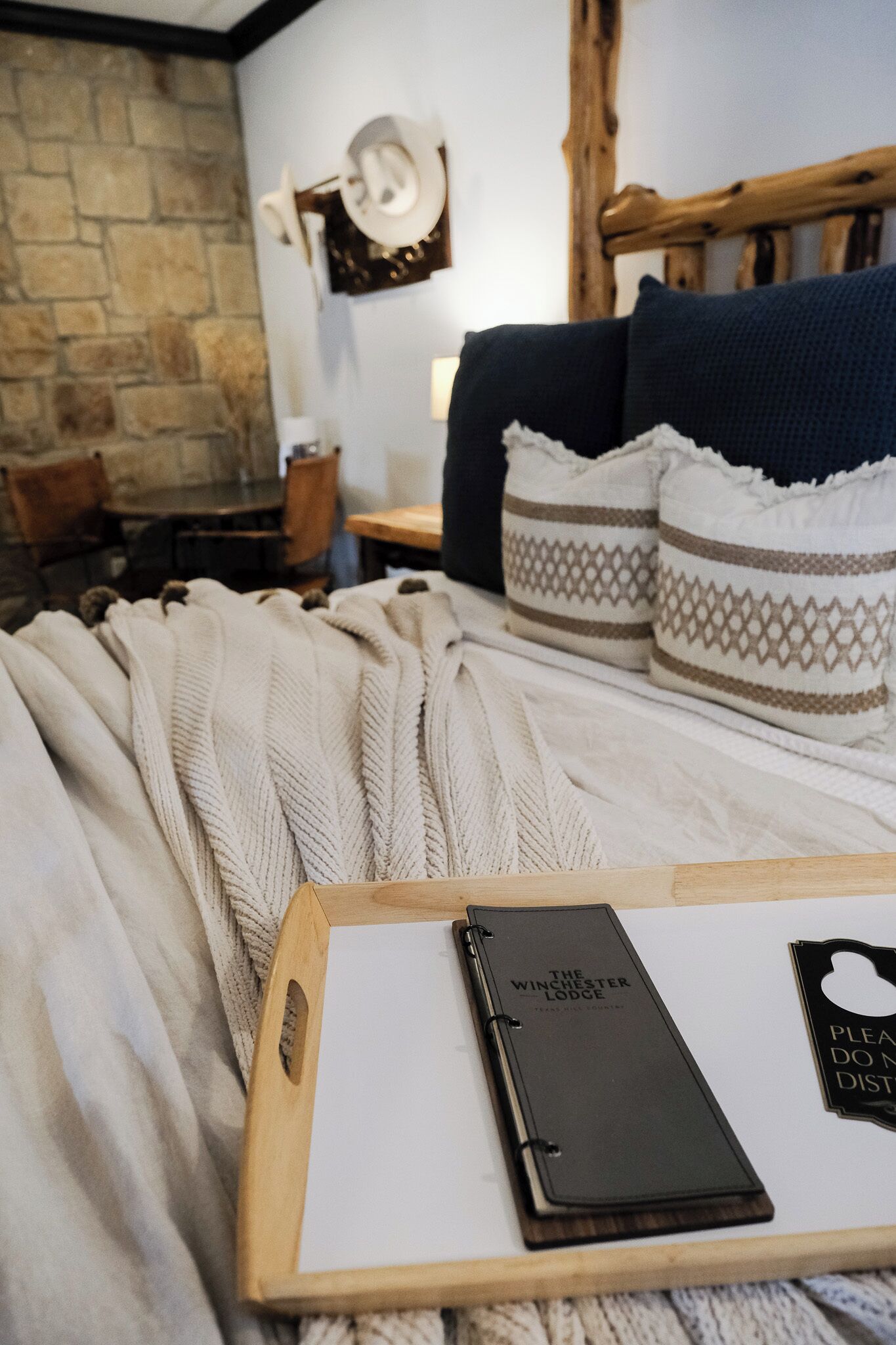 Una cama bien hecha con una manta blanca de punto, un cabecero de madera y almohadas estampadas. Una bandeja de madera sostiene un cuaderno y un bolígrafo.