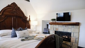 Un dormitorio con una cama grande, una chimenea de piedra, un televisor colocado sobre la chimenea y una mesita de noche con una lámpara.