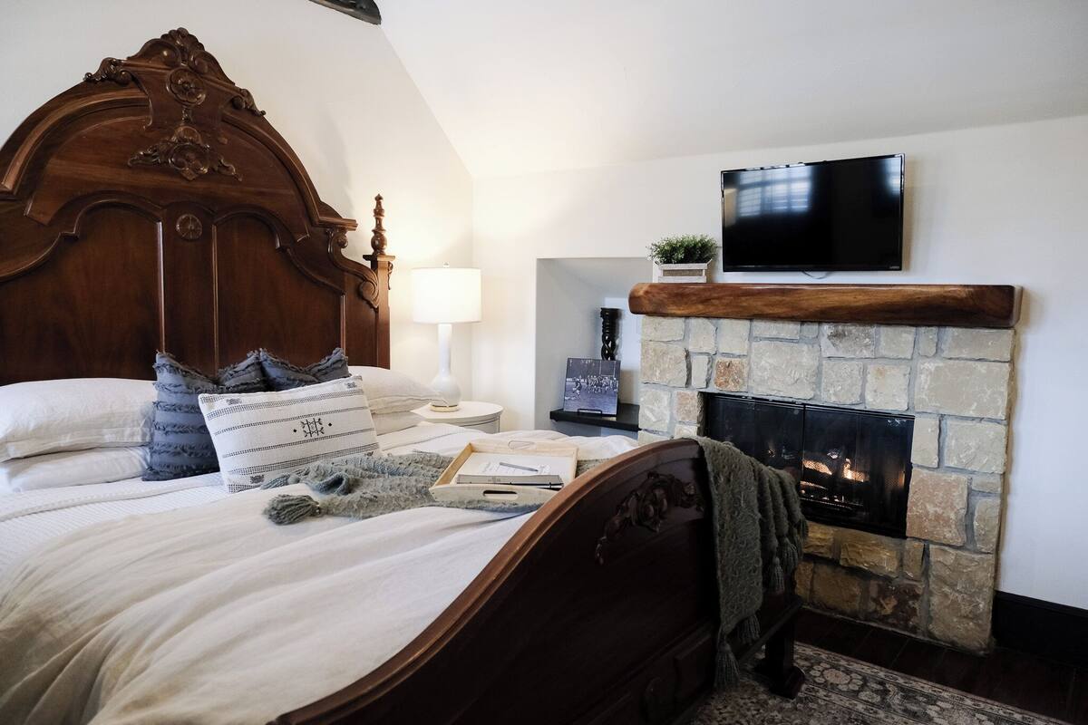Une chambre avec un grand lit, une cheminée en pierre, un téléviseur fixé au-dessus de la cheminée et une table de chevet avec une lampe.