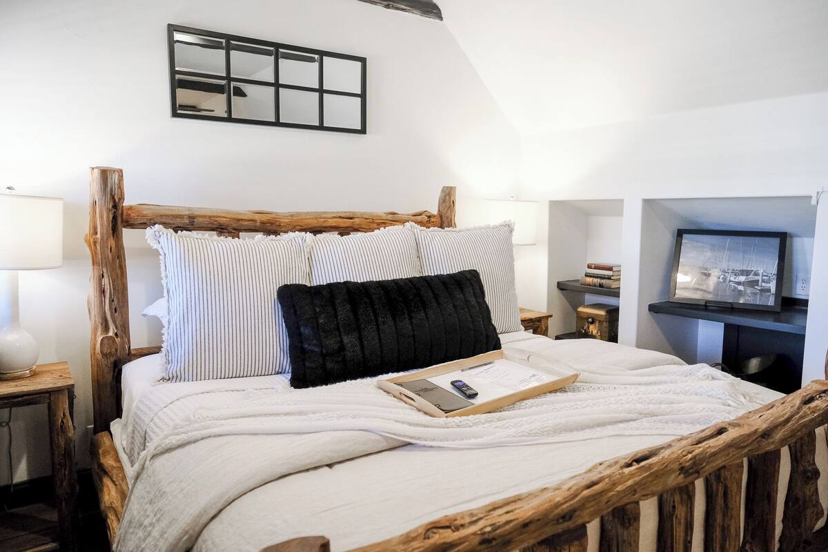 Un lit avec un cadre en bois, une literie blanche, des oreillers à rayures, un oreiller noir, un plateau avec un livre et une télécommande, une lampe, un tableau encadré et un téléviseur posé sur un meuble.