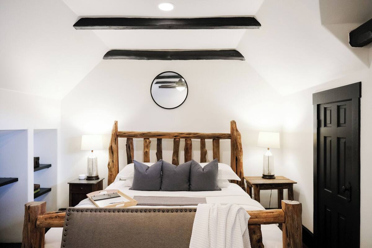 Une chambre à coucher avec un lit en bois, deux tables de chevet avec des lampes, un miroir au mur et une porte noire.