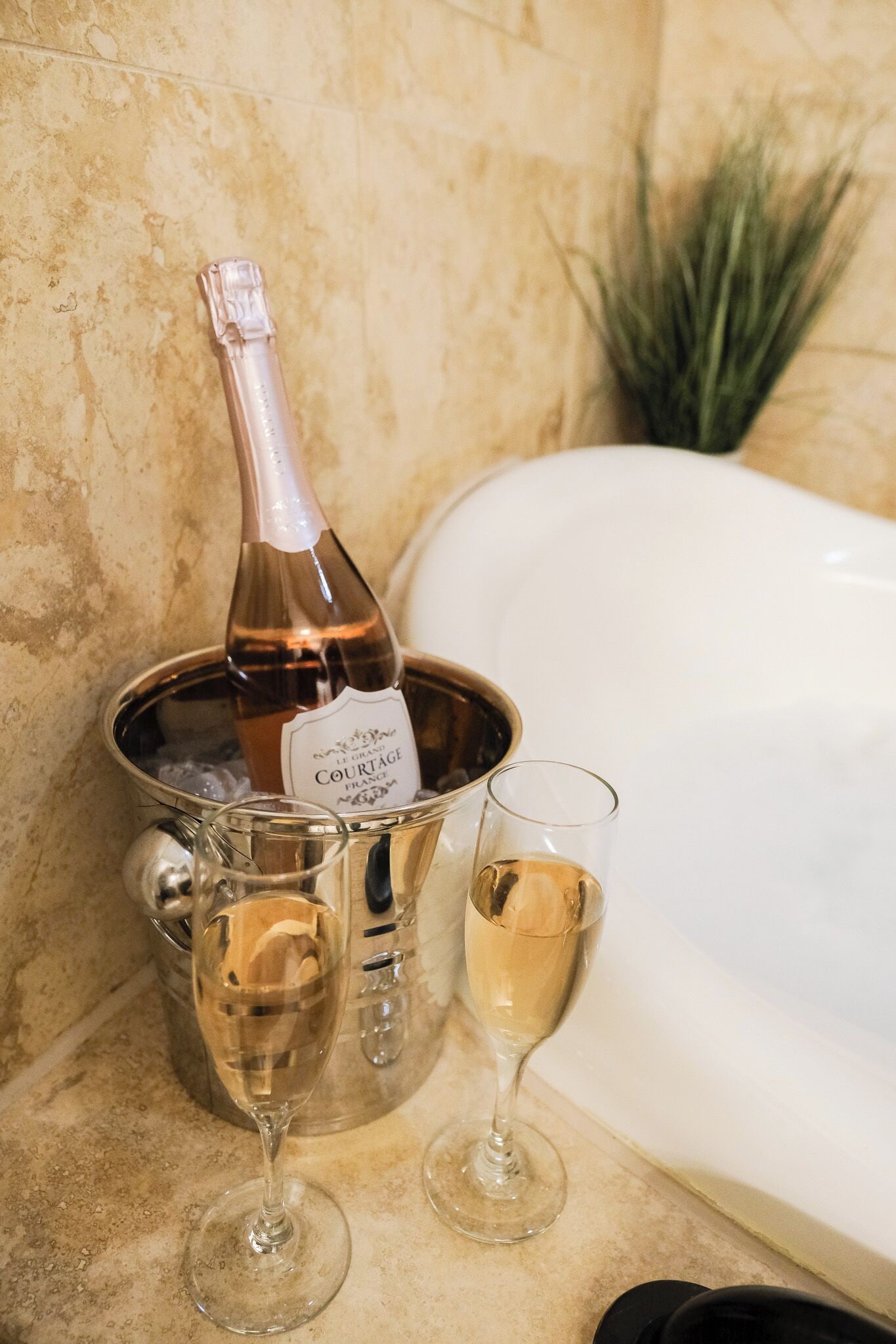 Eine Flasche Courtage Champagner in einem Metallkübel mit zwei Gläsern, eines gefüllt, eines leer, auf einer Marmorarbeitsplatte neben einer Badewanne.