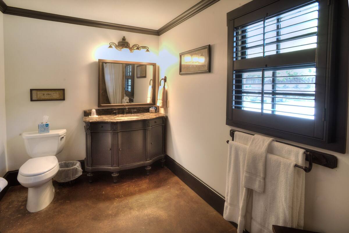 Une salle de bain équipée de toilettes, d’un meuble-lavabo, d’un miroir et d’une fenêtre avec des stores.