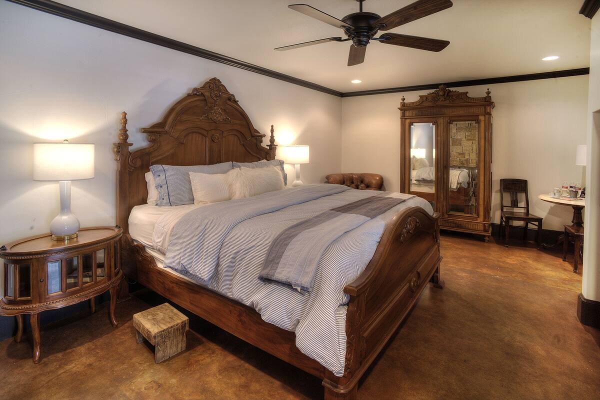 Une chambre à coucher avec un lit en bois, deux lampes de chevet, une table de chevet en bois, un petit tabouret, un ventilateur de plafond, un grand miroir et une chaise en bois.