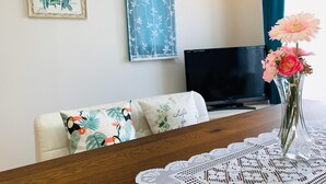 Ein Wohnzimmer mit einer Couch, einem Fernseher, einem Tisch mit Spitzenbettdecke und einer Vase mit Blumen.