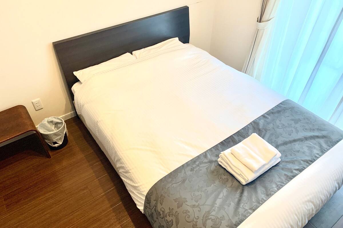Una cama bien hecha con ropa de cama blanca y gris, una mesita de noche pequeña y un cubo de basura.