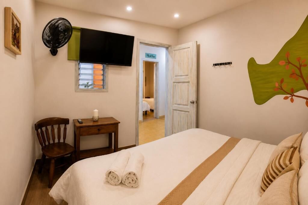 A hotel room with a bed, a chair, a small table with a remote control, a wall-mounted TV, and a door leading to another room.