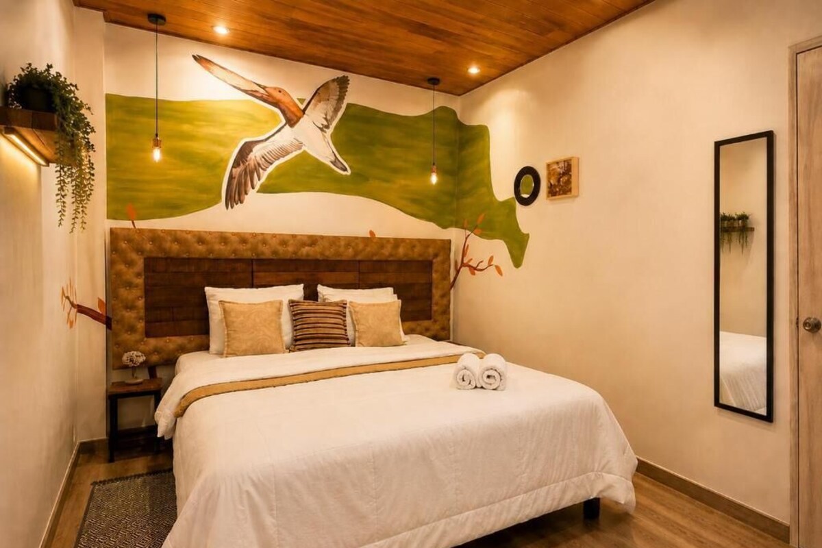 Un dormitorio con una cama grande, cabecera de madera, un mural en la pared, una mesita de noche y un espejo.