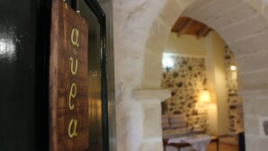 A wooden door with the letters "aqua" carved into it, an arched stone doorway, and a room with a stone wall and a table with a white cloth. - Roes apartments (Faistos)