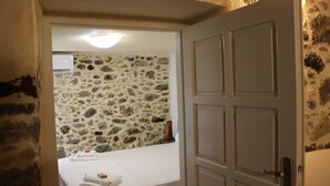 A room with a stone wall, a bed with white linens, and a door with a handle. - Roes apartments (Faistos)