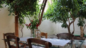 An outdoor dining area with a glass table and wooden chairs, surrounded by trees and a white wall. - Roes apartments (Faistos)