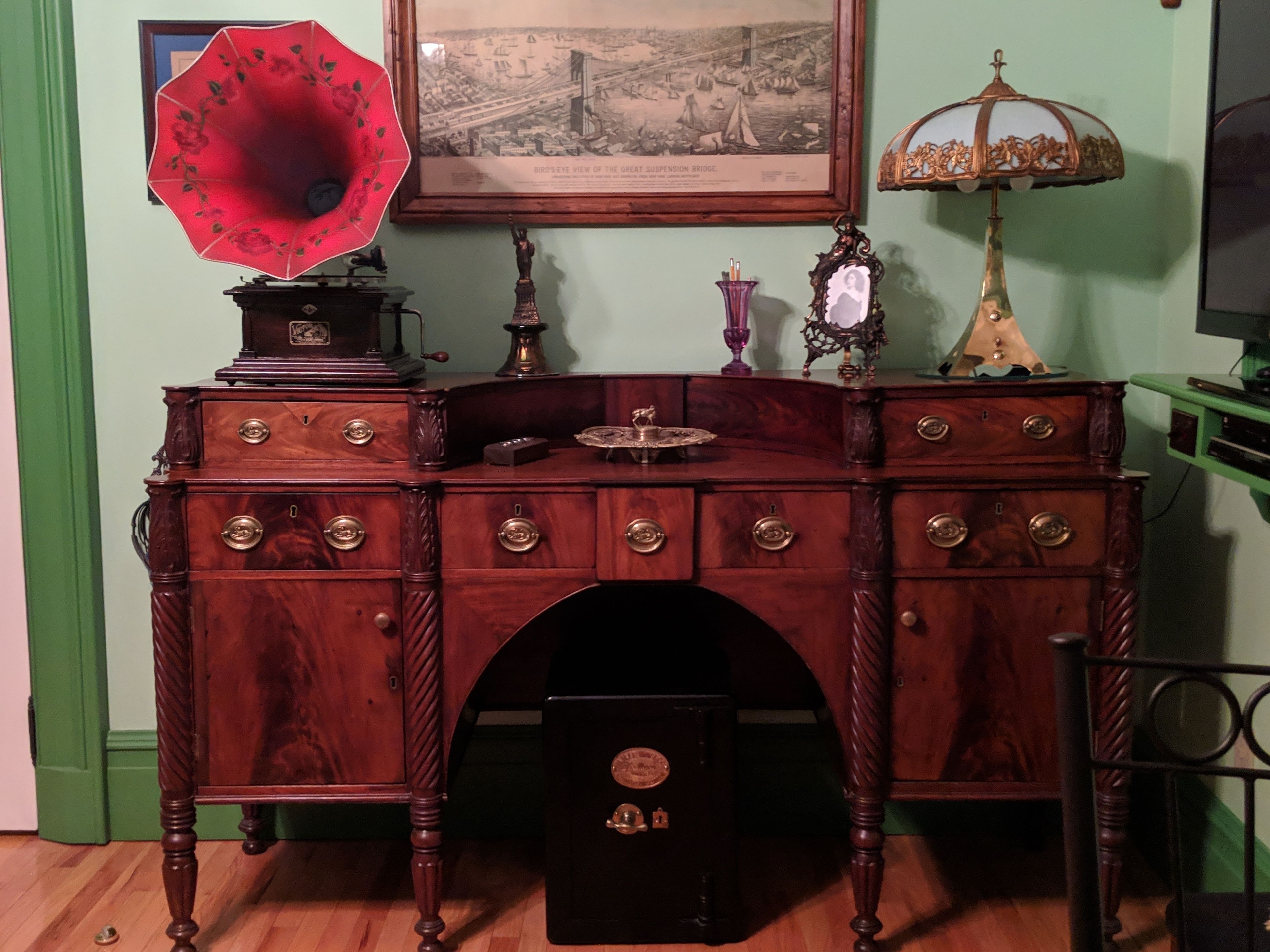 Un bureau en bois ancien, doté de tiroirs, un phonographe, une vue de la ville encadrée et une lampe.