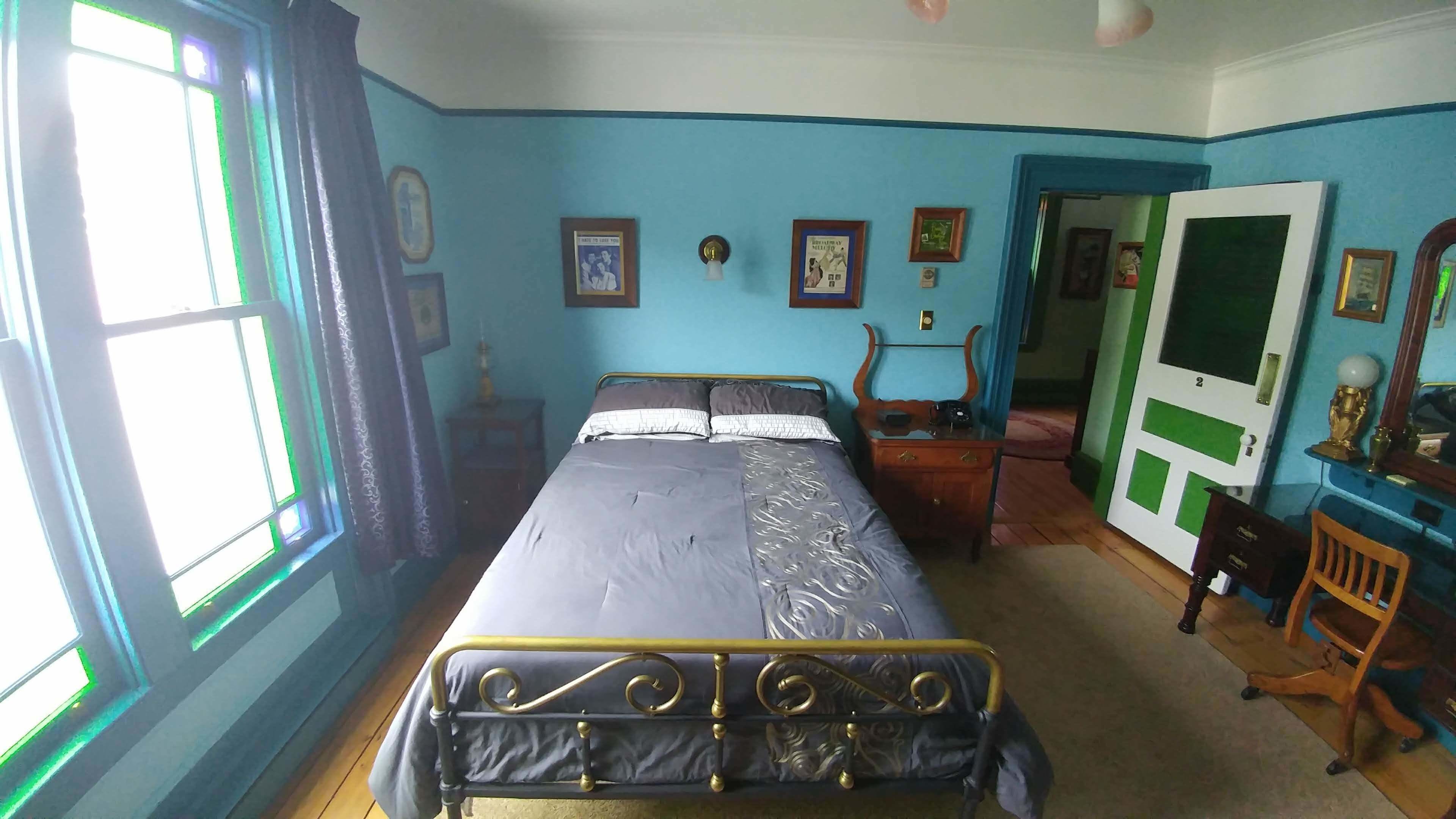 Una habitación con una cama, un escritorio y una silla. Hay cuadros en la pared y una puerta con un diseño verde y blanco.