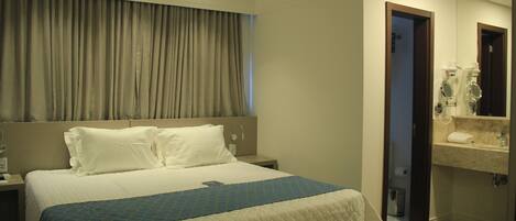 Habitación de hotel con una cama grande, una mesita de noche, un baño con espejo y lavamanos, y una cortina que cubre la ventana.
