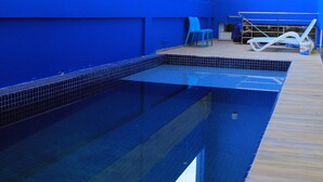 An indoor swimming pool with blue walls and a tiled floor, surrounded by lounge chairs and a small seating area. - Blue Open Hotel (Erechim)