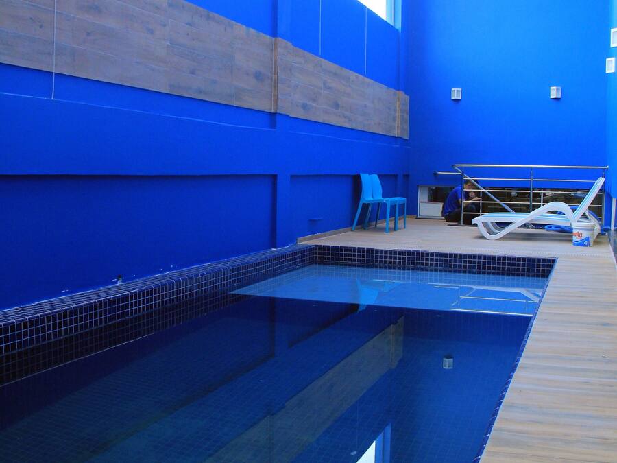 Una piscina interior con paredes azules y suelo de baldosas, rodeada de sillones de descanso y una pequeña zona de estar.