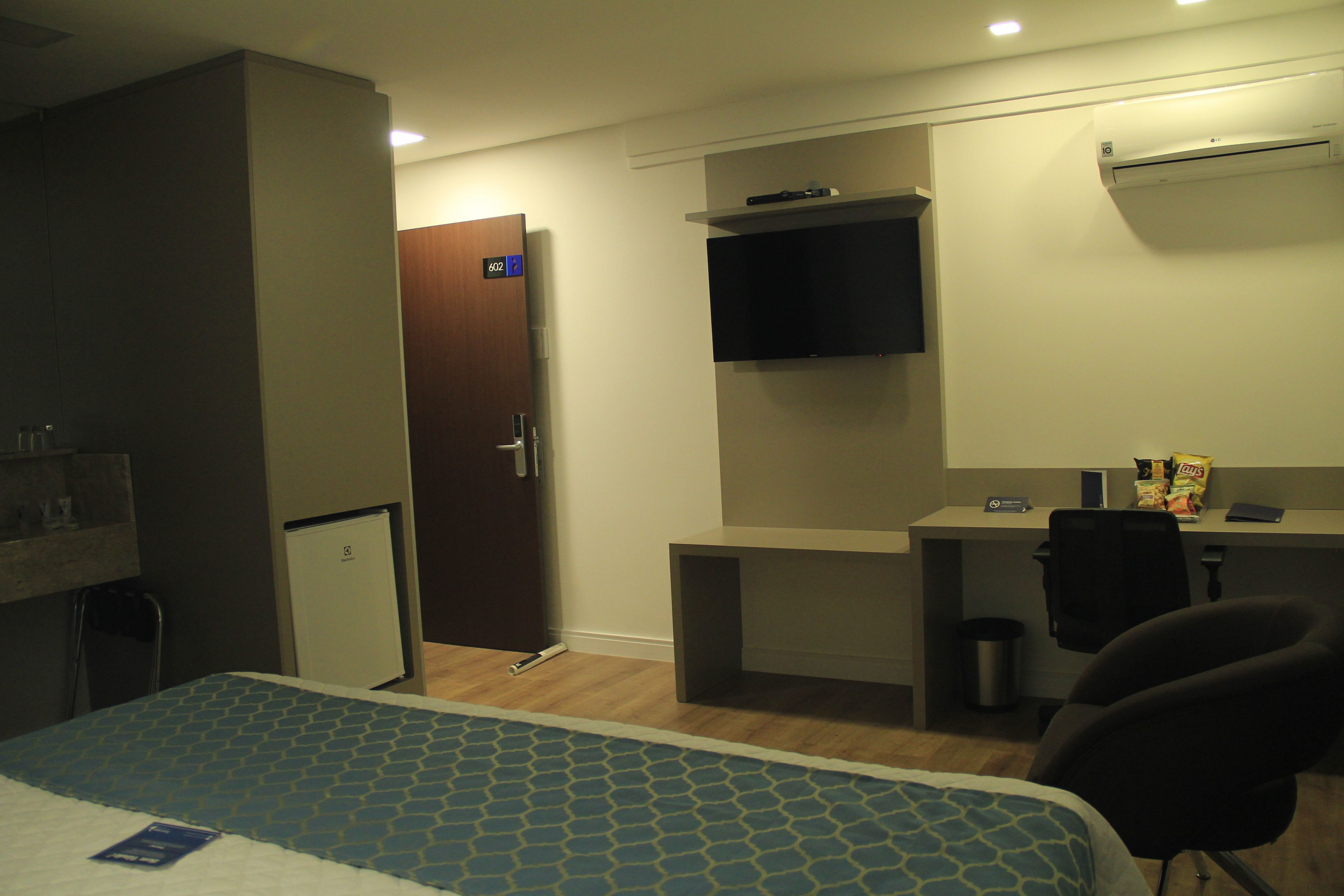 Habitación de hotel con cama, escritorio, silla, televisor y puerta.