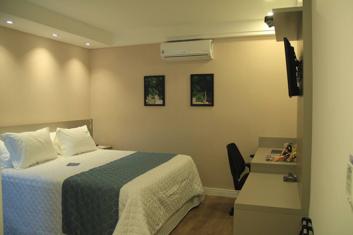A hotel room with a bed, a desk, an air conditioner, and two framed pictures on the wall.