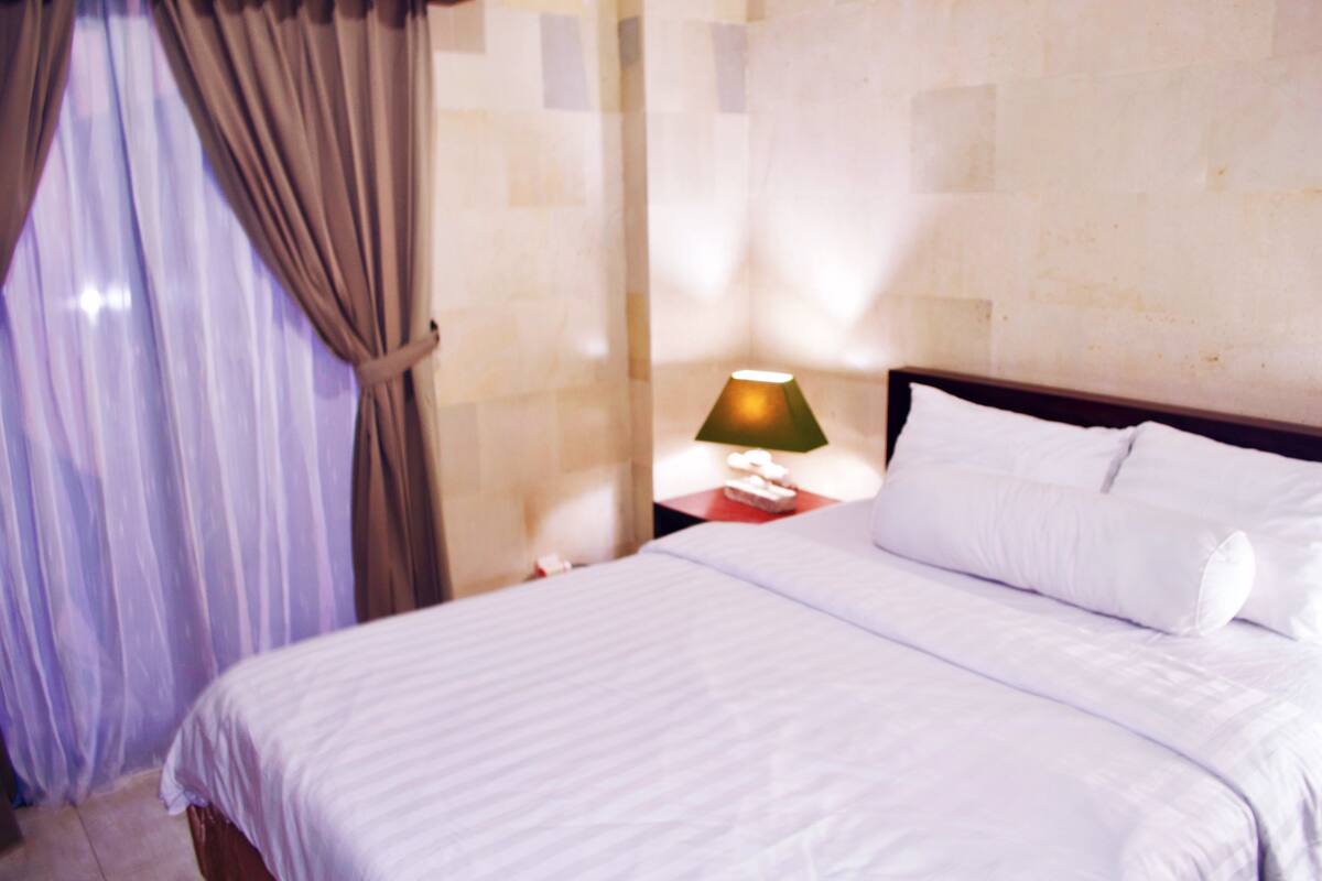 A hotel room with a bed, curtains, a nightstand with a lamp, and a wall with a textured pattern.