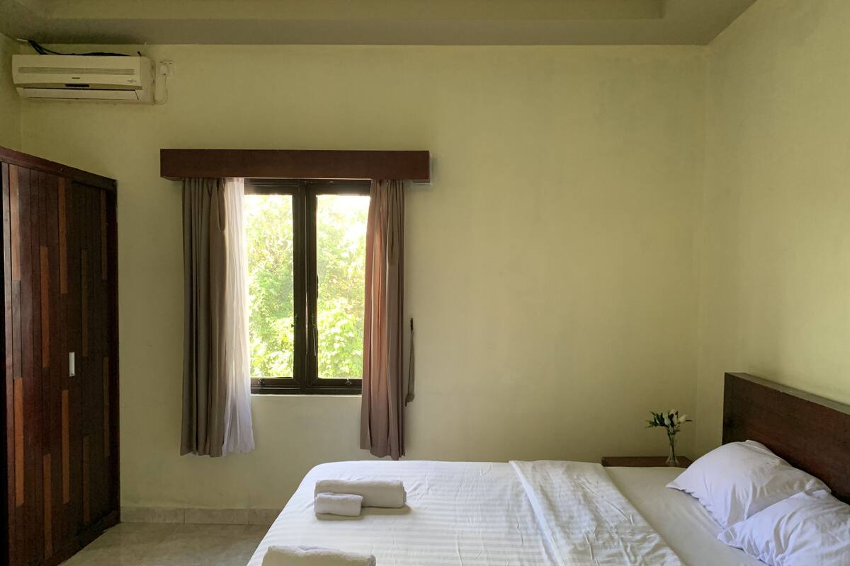 A bedroom with a bed, a window with curtains, a wooden wardrobe, and an air conditioning unit.