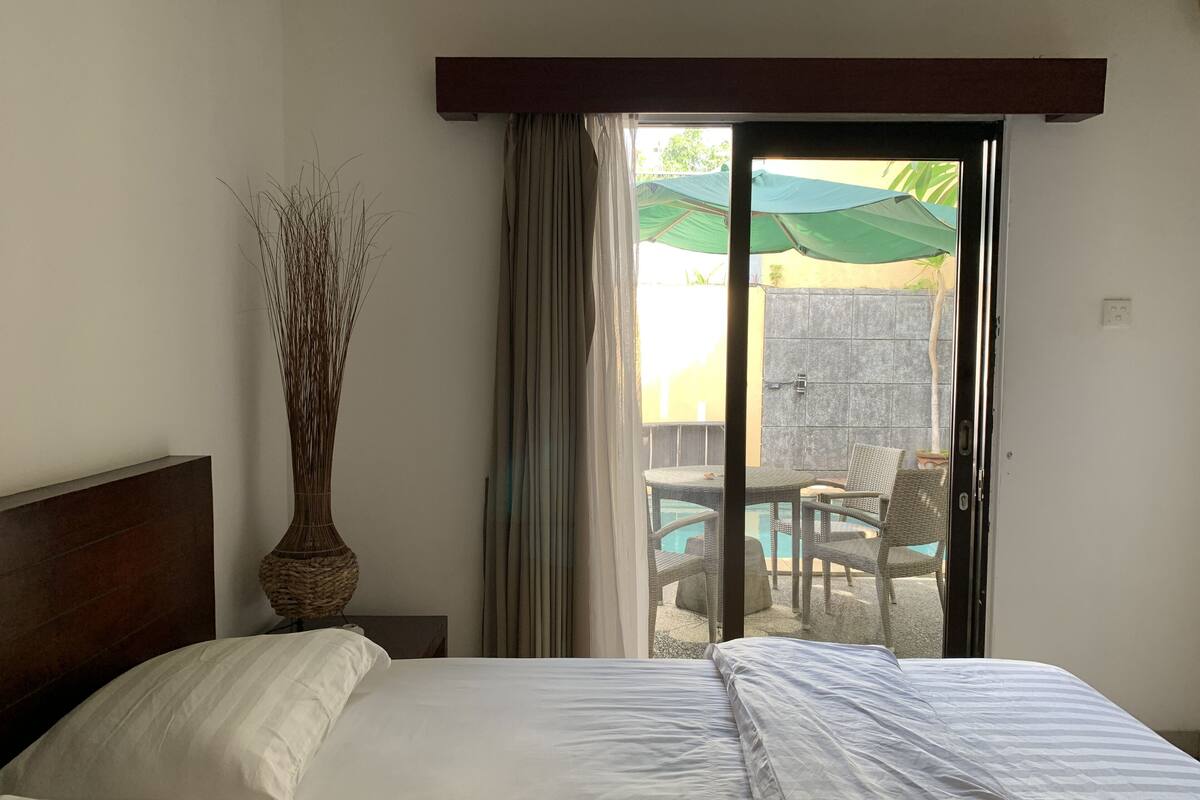A hotel room with a bed, a vase, and a view of an outdoor area with a pool and chairs.