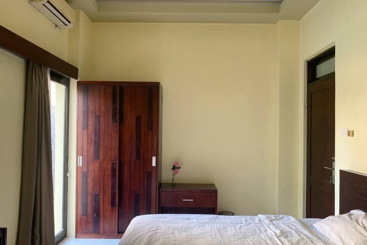 A hotel room with a large bed, a wooden wardrobe, a small desk with a vase, and a window with curtains.