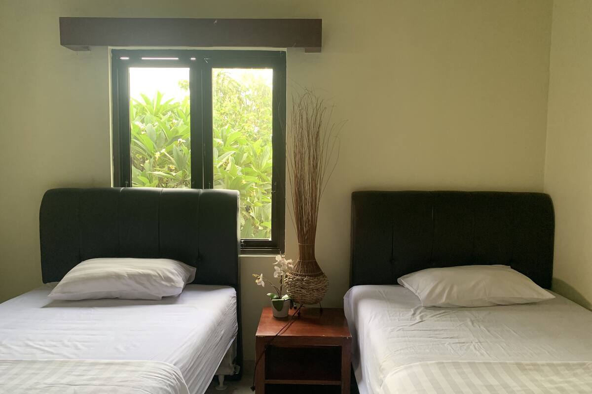 A room with two single beds, a small wooden nightstand, a window with a view of greenery, and a vase with dried plants.