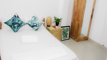 A neatly made bed with white linens, a wooden nightstand with a lamp, a framed plant, and a wooden wardrobe.