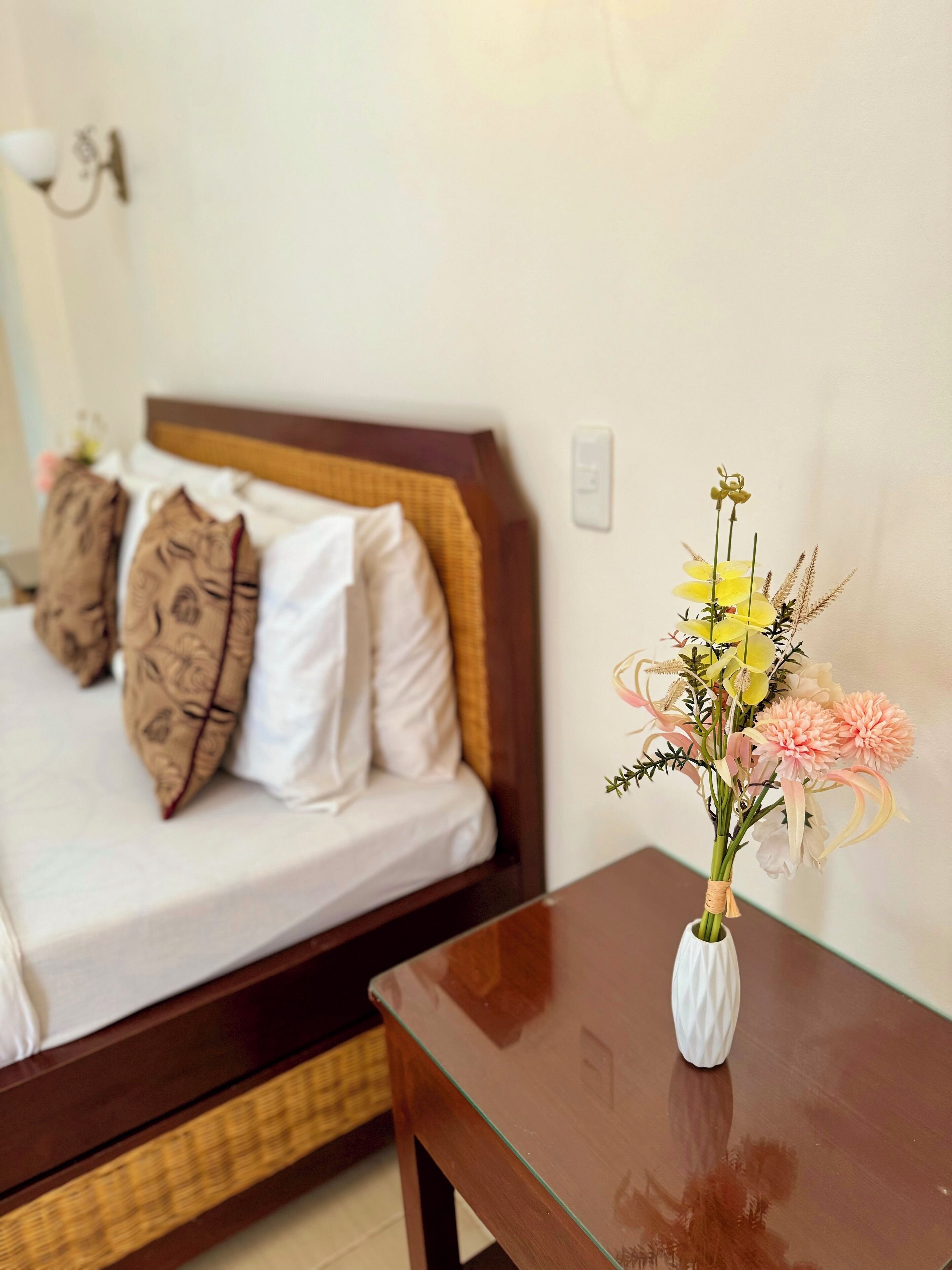 Et hotelværelse med en seng, puder, et natbord med en vase blomster og en lampe på væggen.