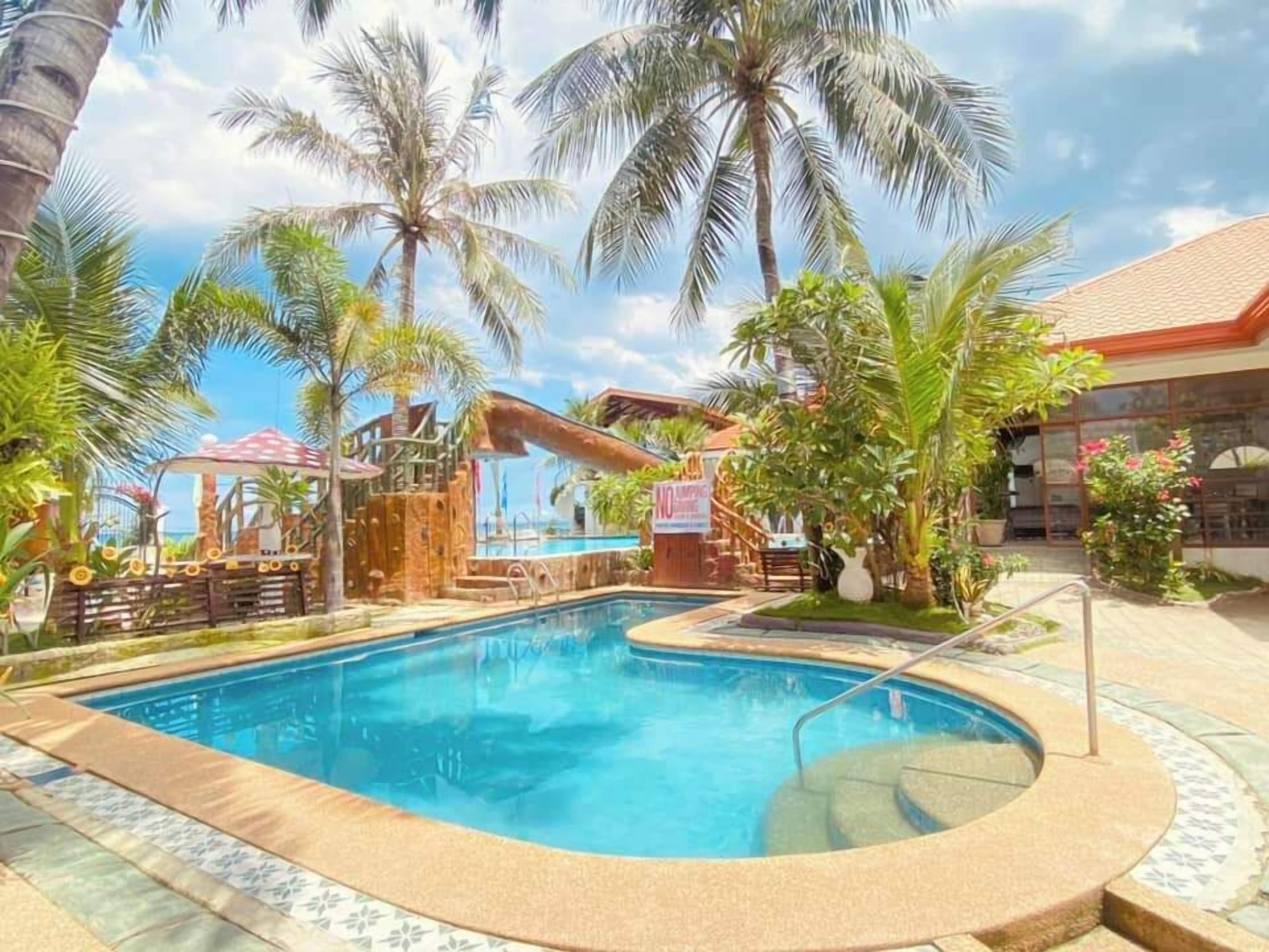 Un espace piscine doté d’une terrasse en bois, de palmiers et d’un bâtiment au toit rouge.