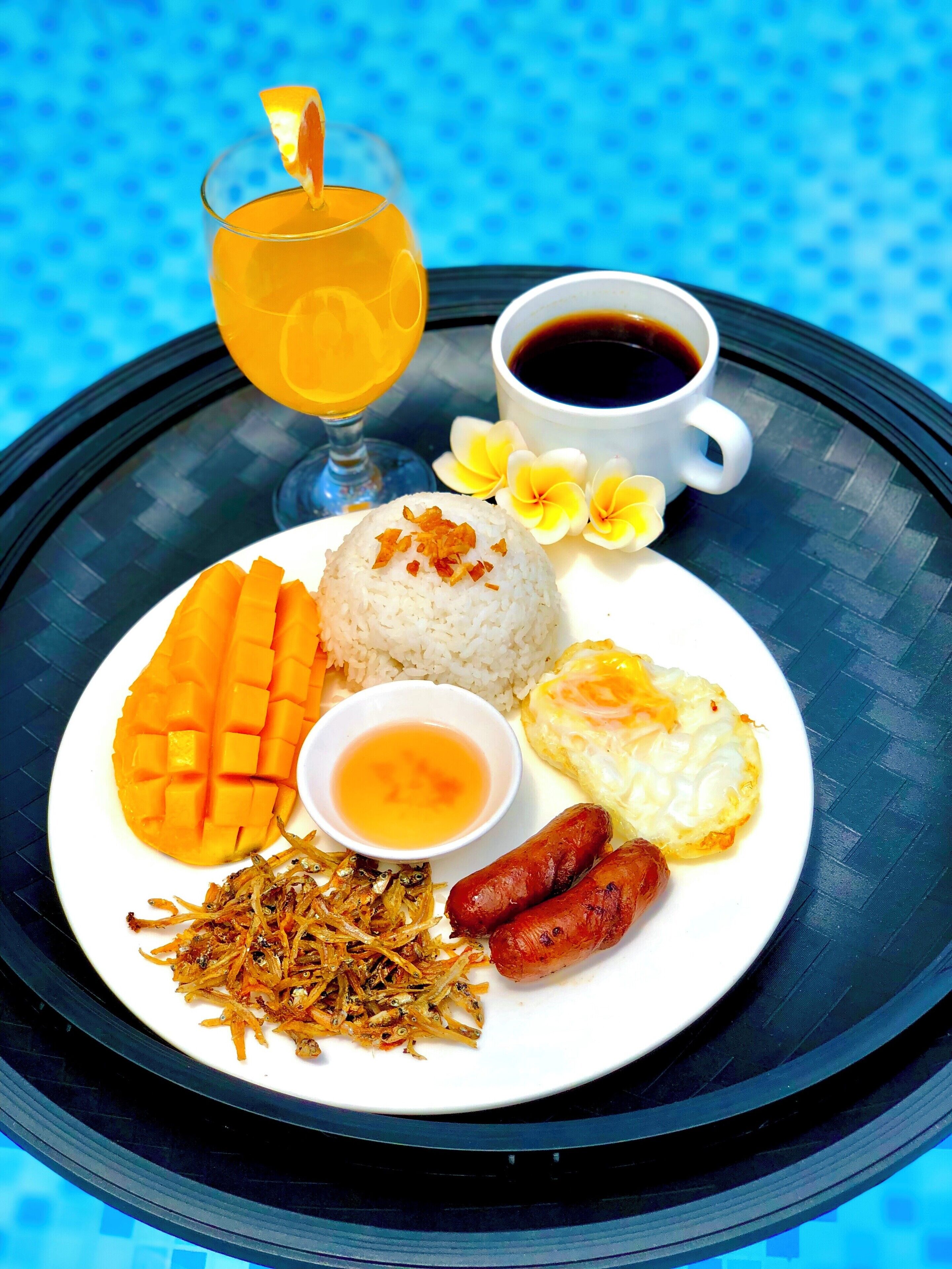 Un plato de comida que incluye arroz, mango, cebolletas fritas, salchichas y un huevo frito, con un vaso de jugo de naranja y una taza de café sobre una bandeja negra.