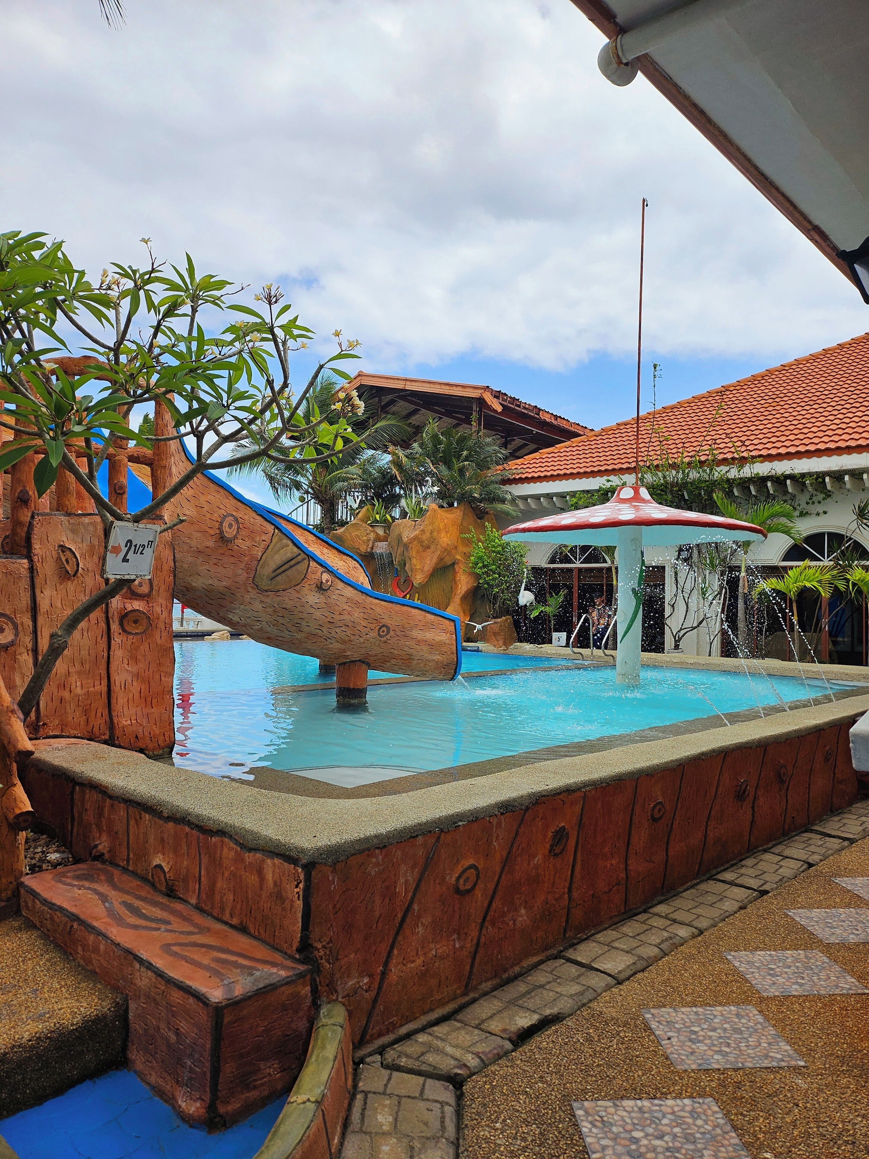 Zona de piscina con un tobogán de madera, una fuente y una zona de estar cubierta.