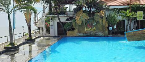 A swimming pool with a hot tub, surrounded by palm trees and a building with a red-tiled roof.