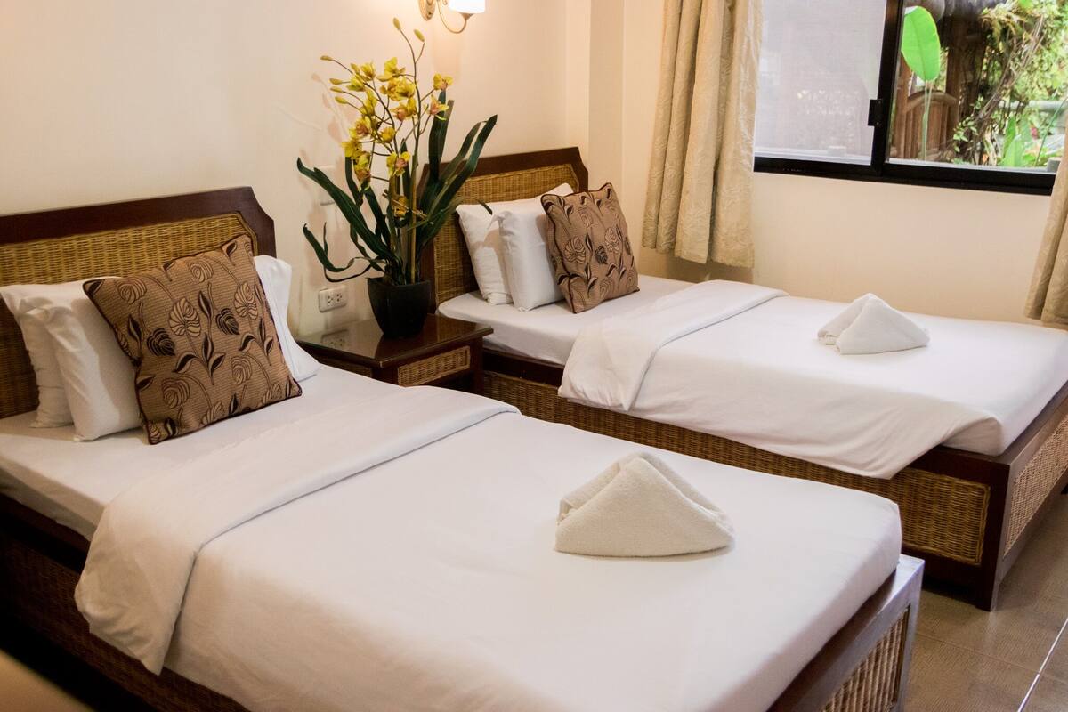 Ein Hotelzimmer mit zwei Betten, jeweils mit weißer Bettwäsche und dekorativen Kissen. Auf einem Nachttisch zwischen den Betten steht ein Topf mit gelben Blumen.