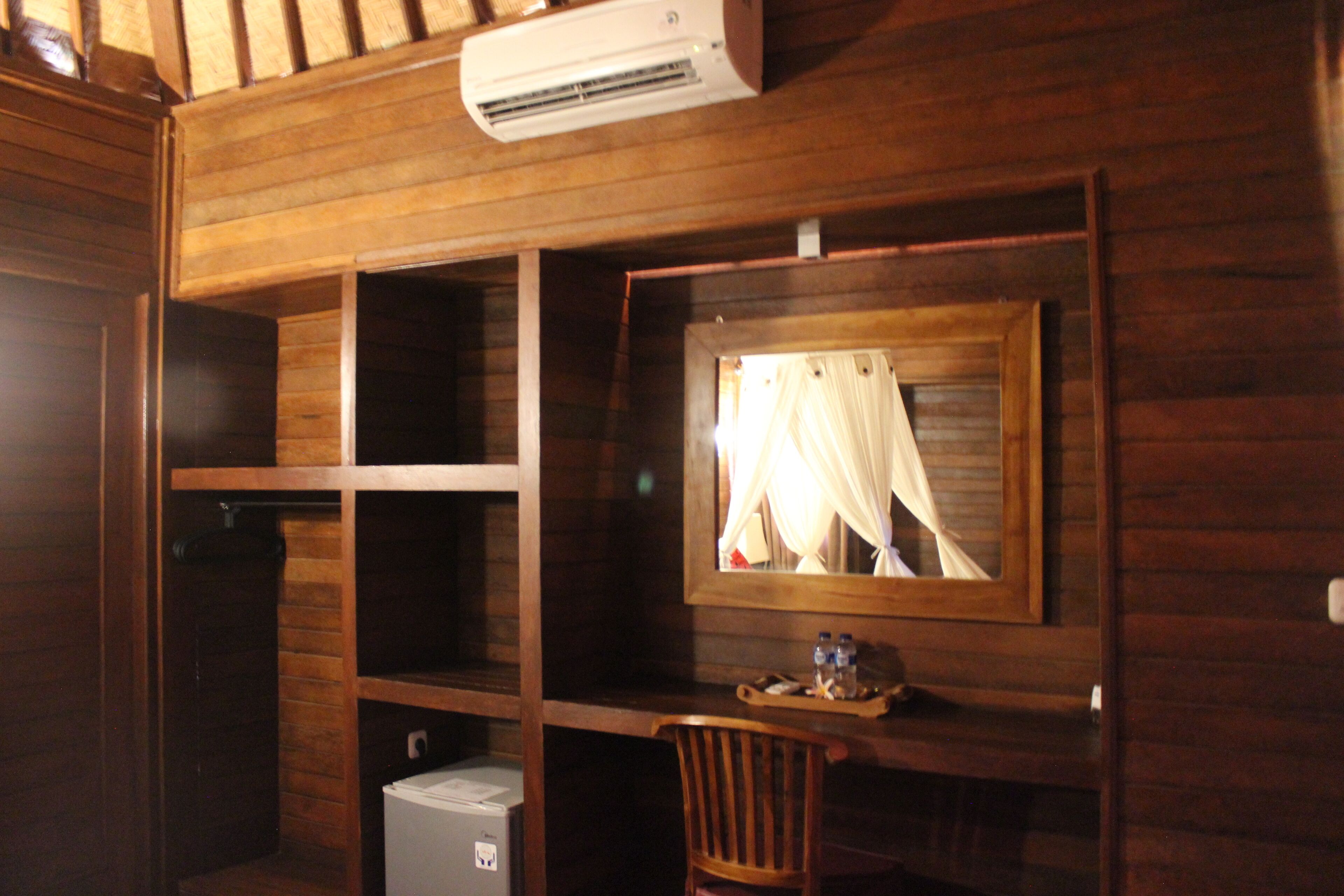 Une pièce en bois avec un bureau, une chaise et un miroir. Il y a un climatiseur au-dessus du bureau et un petit réfrigérateur en dessous.