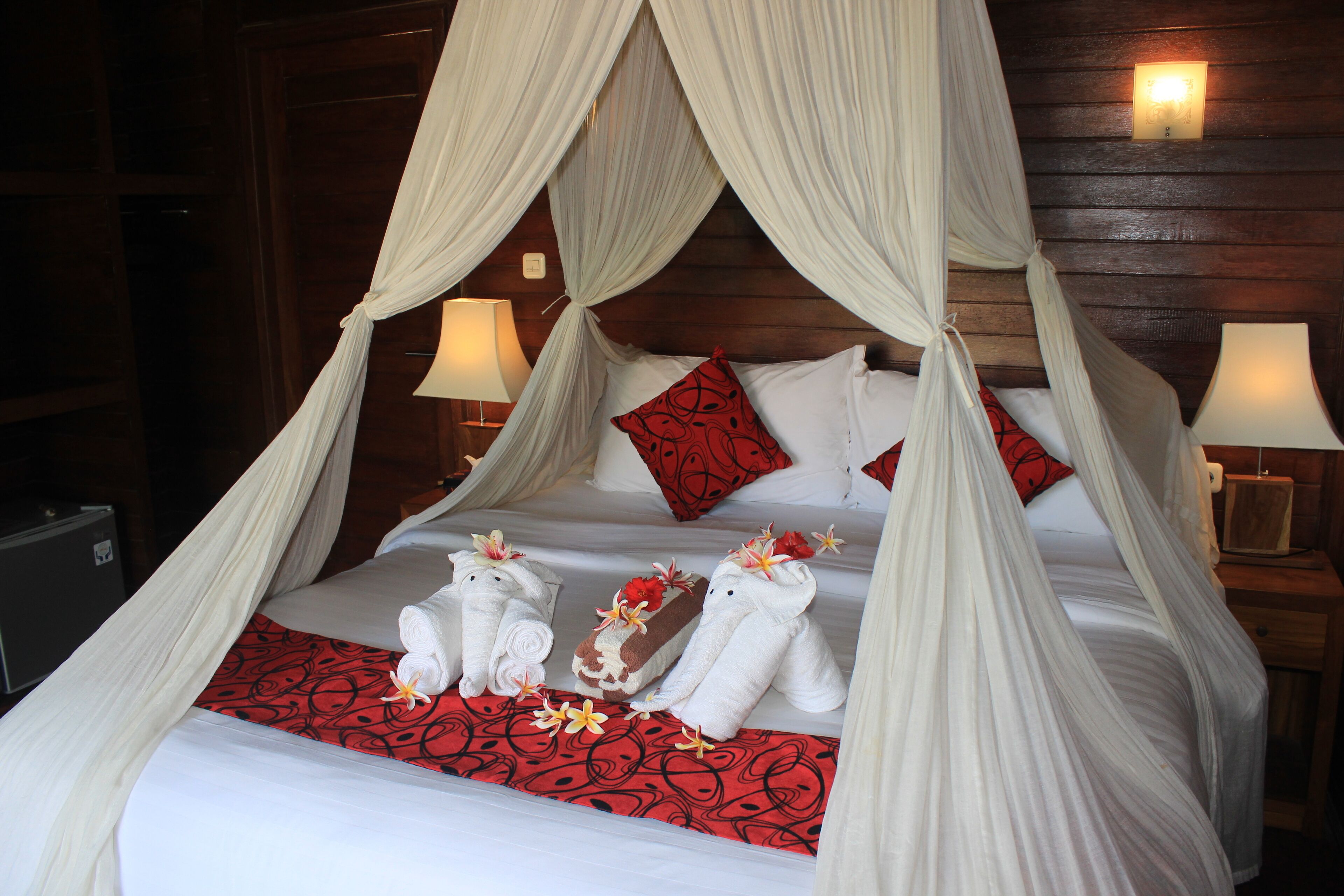 A four-poster canopy bed with white linens, red and black patterned bedspread, and decorative towels with flowers.