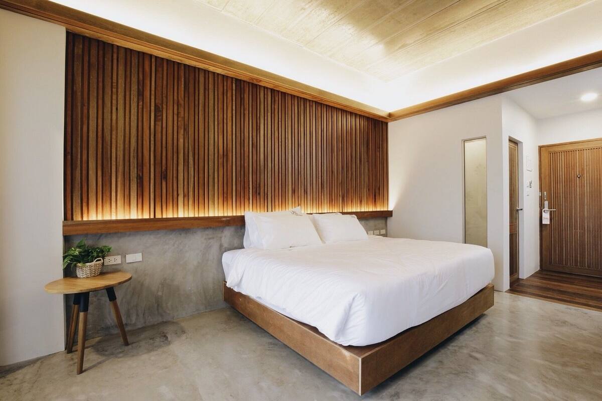 Un dormitorio moderno con cabecera de madera, una cama con ropa blanca, una mesita redonda con una planta y una puerta de madera.